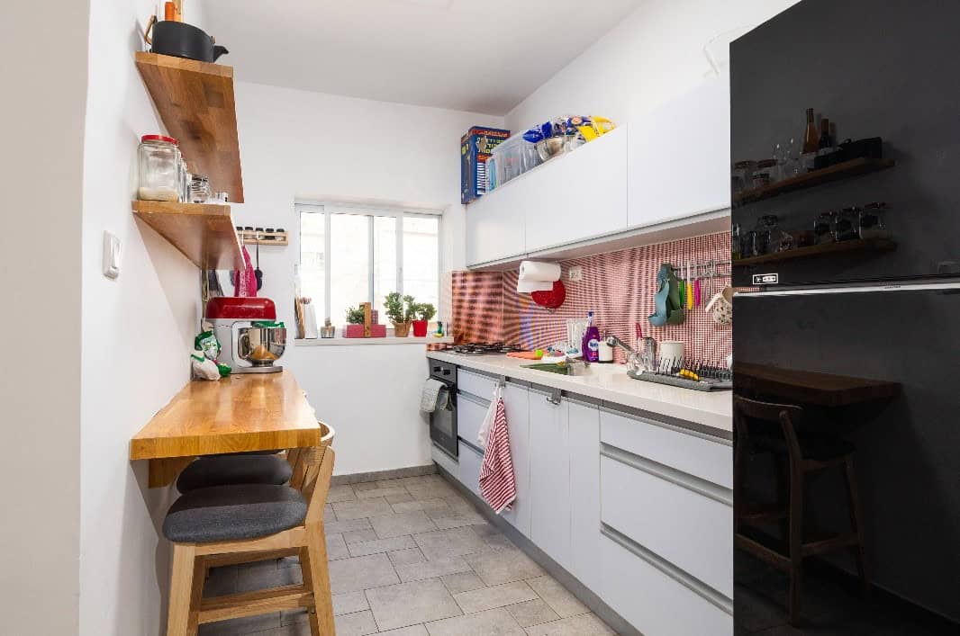 Narrow, bright kitchen with white cabinets, red checkered backsplash, and a wooden breakfast bar with stools on the left.