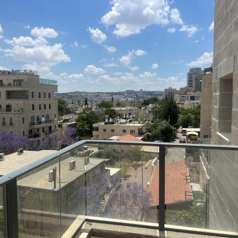 Balcony railing overlooks a cityscape of beige mid-rise buildings and purple flowering trees under a blue sky.