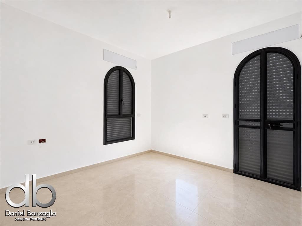 Bright, empty room with white walls, a beige tiled floor, and two tall arched windows/doors with black frames and shutters; a logo sits in the bottom-left corner of the image.