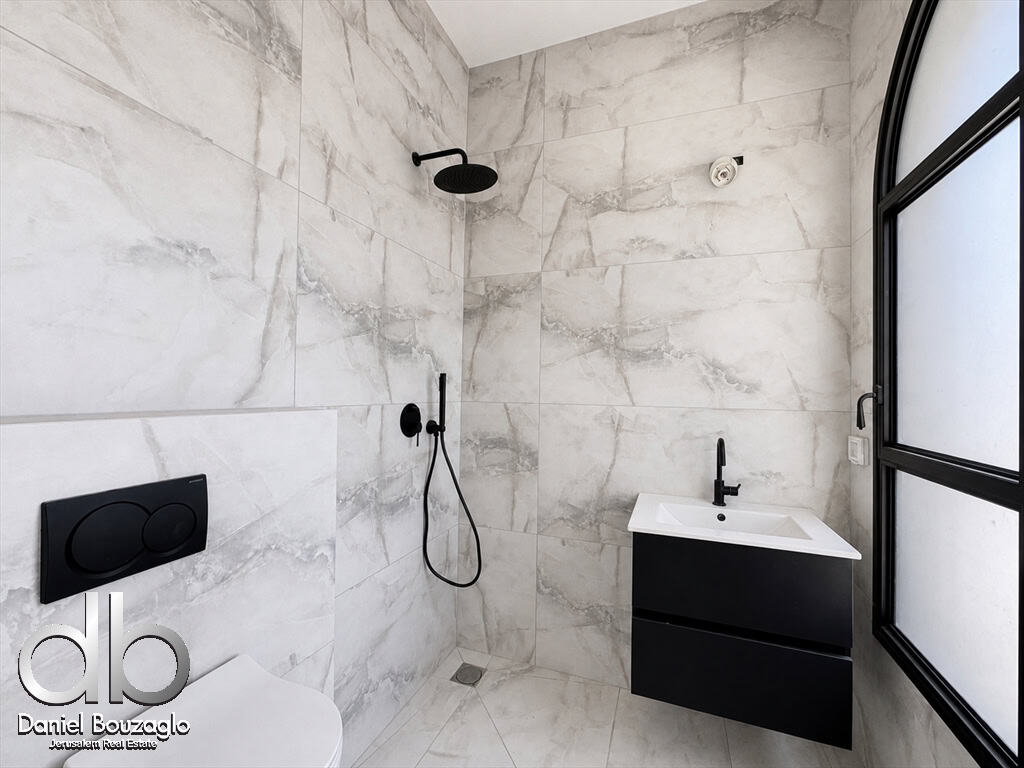 Modern marble bathroom with black fixtures: wall-mounted toilet, rainfall shower, handheld shower, and a black vanity under a large frosted window.