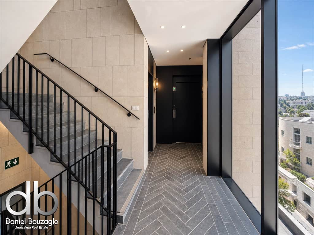 Modern building lobby with gray tile floor, black metal staircase railing, and a large window view of the cityscape on the right side.