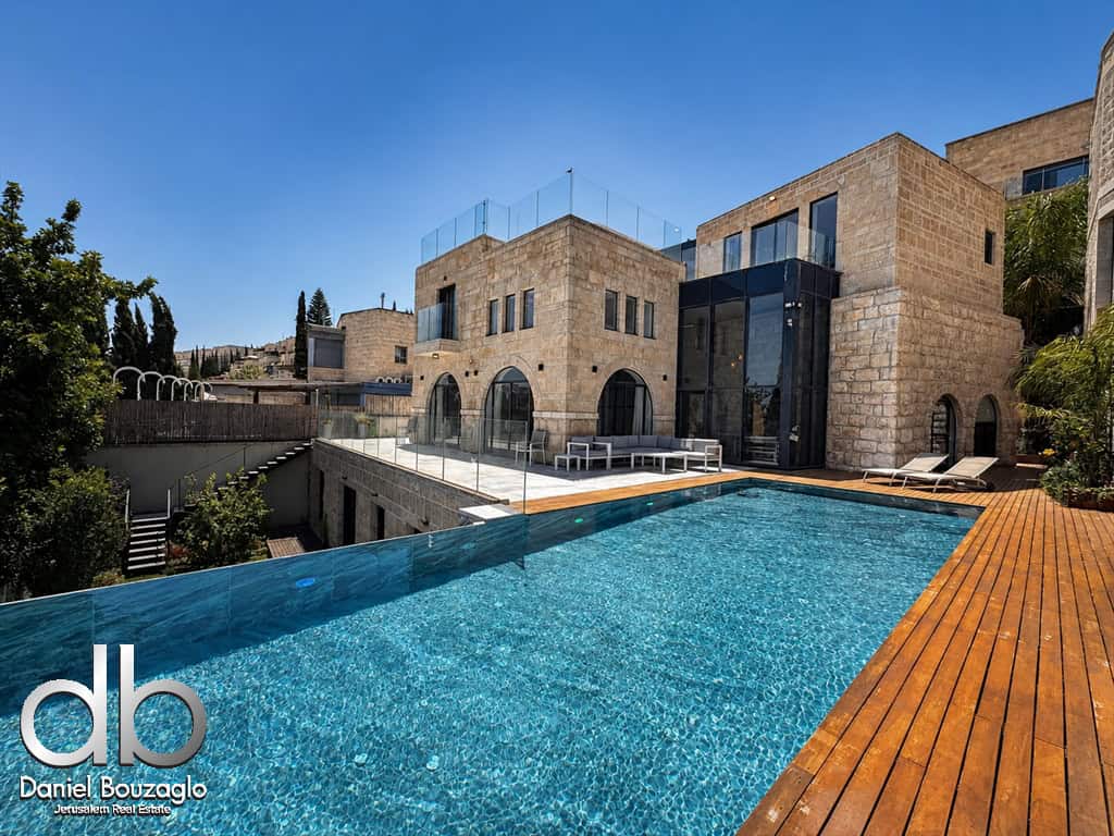 Luxurious stone house with a long wooden deck and an outdoor pool under a clear blue sky.