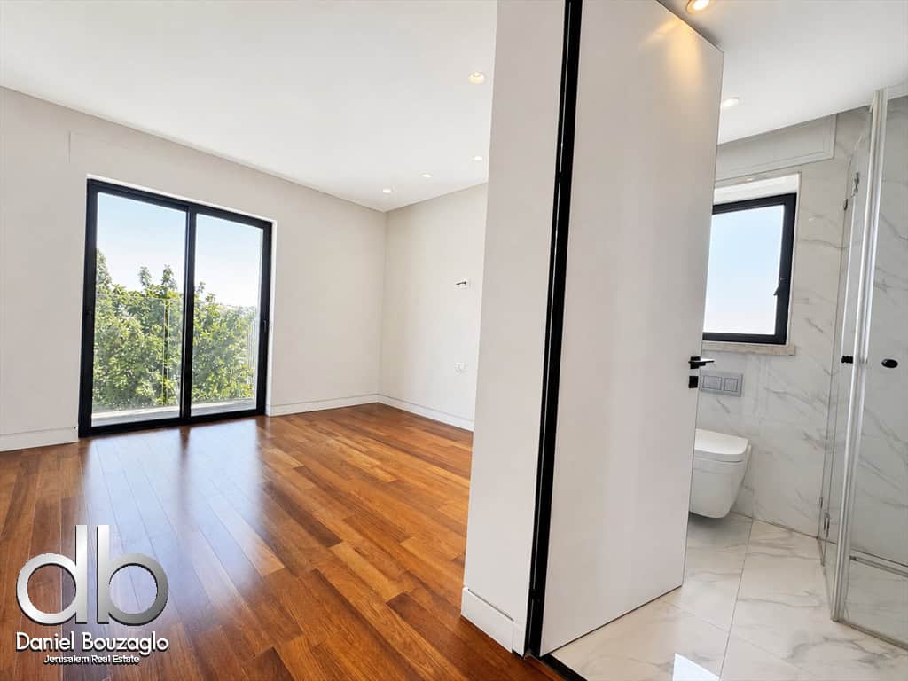 Bright modern living space with warm wood floors, a large sliding glass door to a balcony, and an open view of greenery outside; partial view of a marble-tiled bathroom with a glass shower.