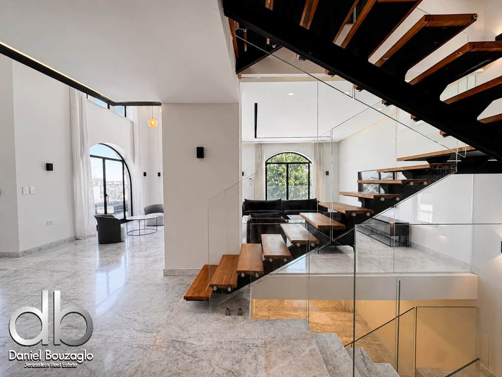 Open-concept living area with a floating wooden staircase, glass railing, white marble floors, and a large arched window overlooking greenery.