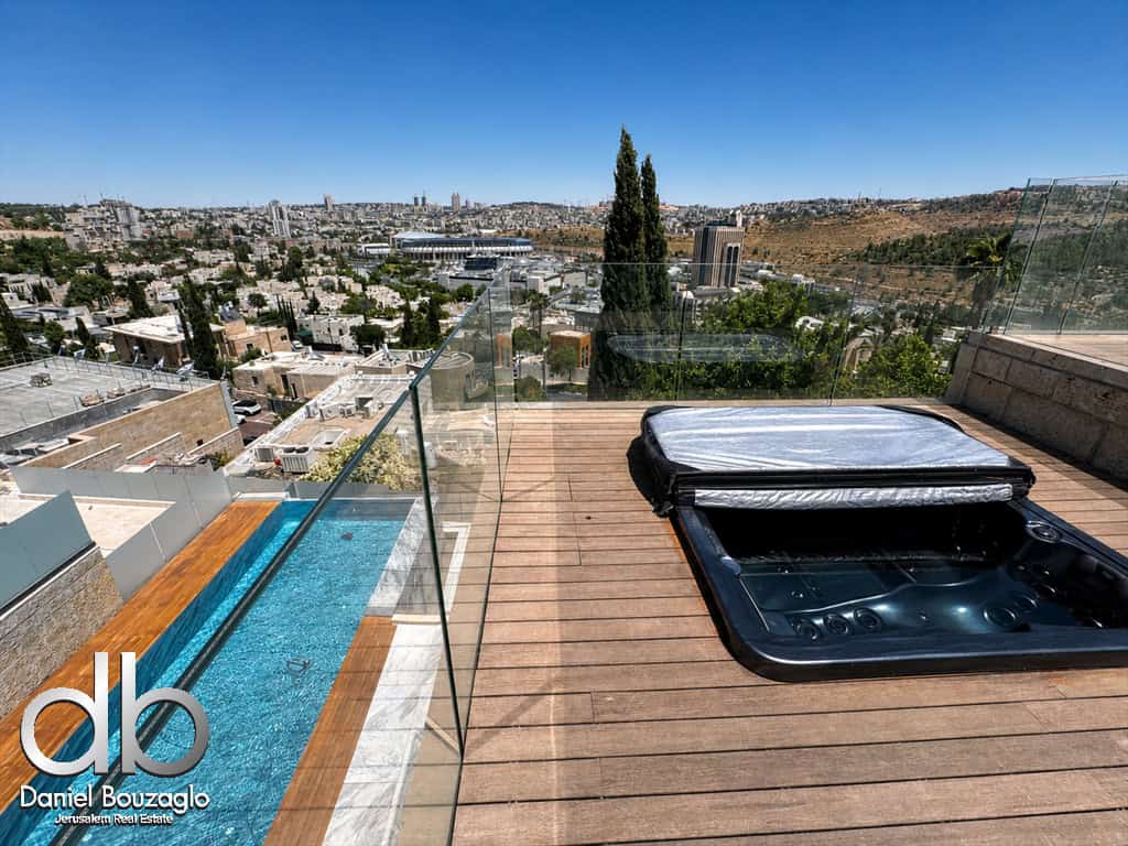 Rooftop deck with a hot tub on wooden flooring, glass railing, and a cityscape view under a clear blue sky; pool to the left.