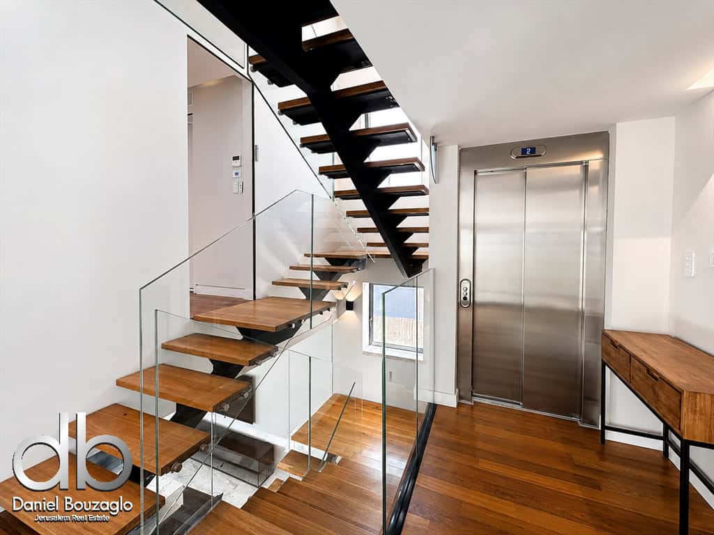 Modern interior with floating wooden stairs and glass railing beside an elevator and a wooden desk.