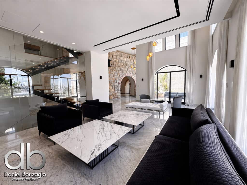 Modern hotel or showroom lobby with marble tables, black couches, and a glass-walled staircase near a stone accent wall and large arched windows.