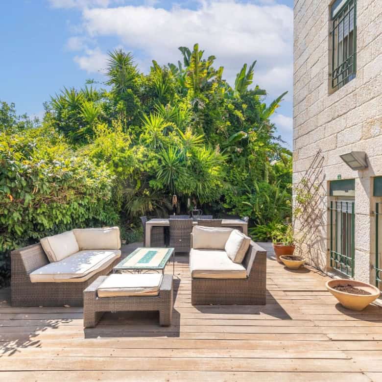 Cozy outdoor wooden deck with wicker furniture and cream cushions, surrounded by lush green plants beside a stone building.