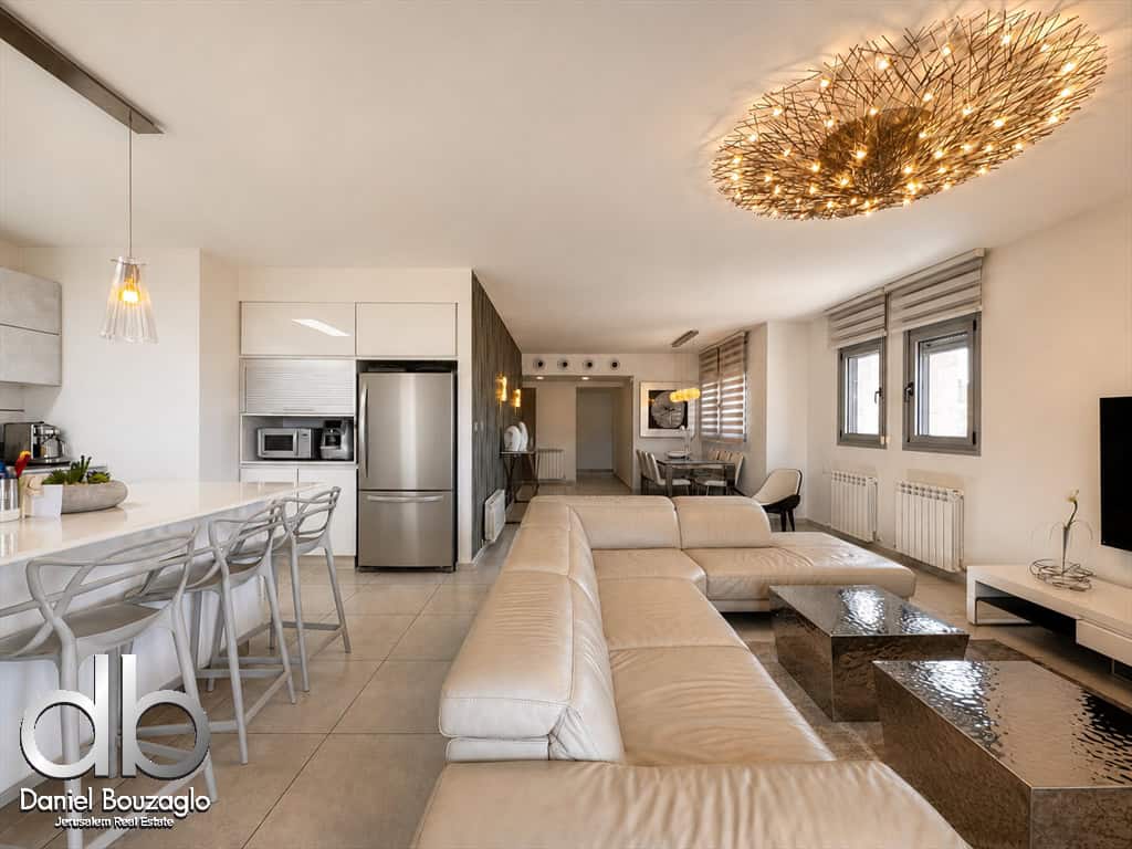 Open-concept living area with a beige leather sectional, two rectangular coffee tables, and a wall-mounted TV in a bright, modern kitchen/dining space. Watermark in lower-left corner reads 'Daniel Bouzaglo'.