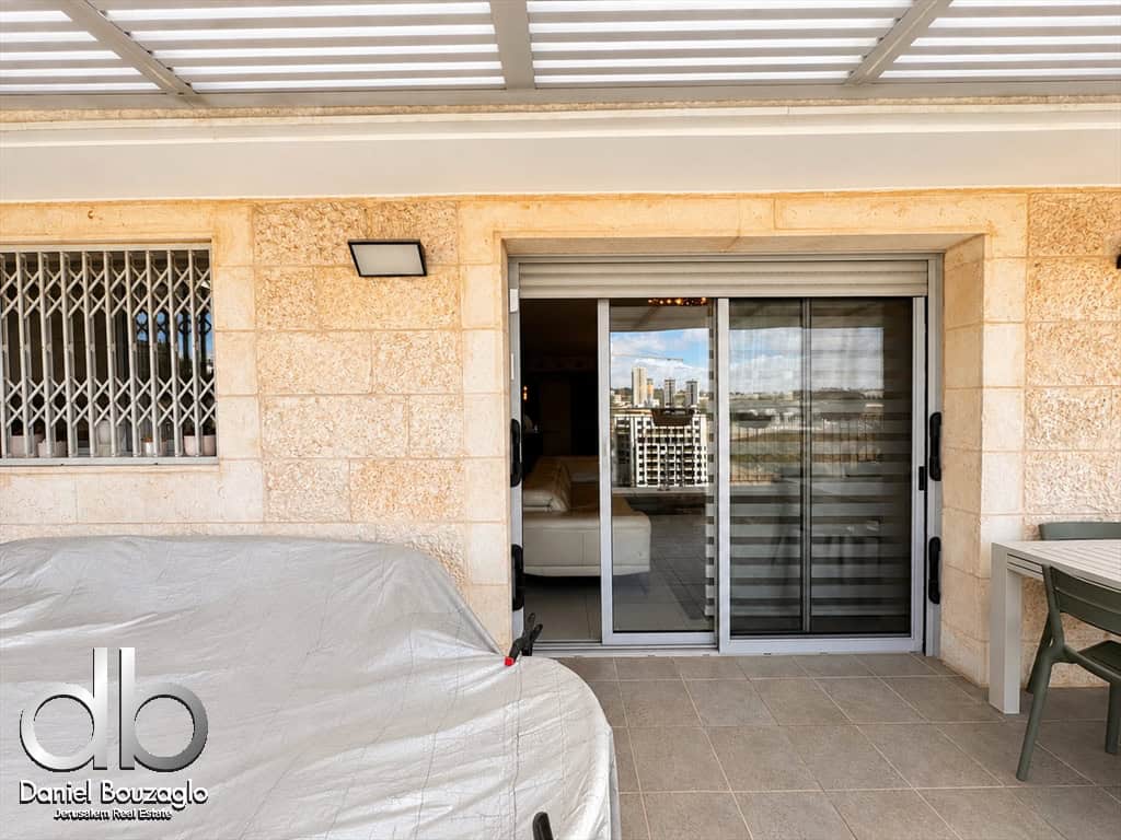 Patio with a sliding glass door opening to a living room, a covered outdoor item in the foreground, and a plastic patio table nearby on a tiled floor.