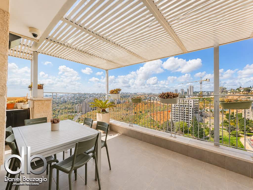 Balcony dining area with a white table and six chairs, potted plants, and city skyline under a blue sky. The space is shaded by a slatted overhead pergola.