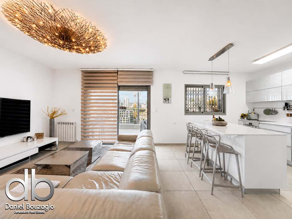 Open-plan living room and kitchen with a beige leather sofa, coffee tables, wall TV, and a white island with bar stools.