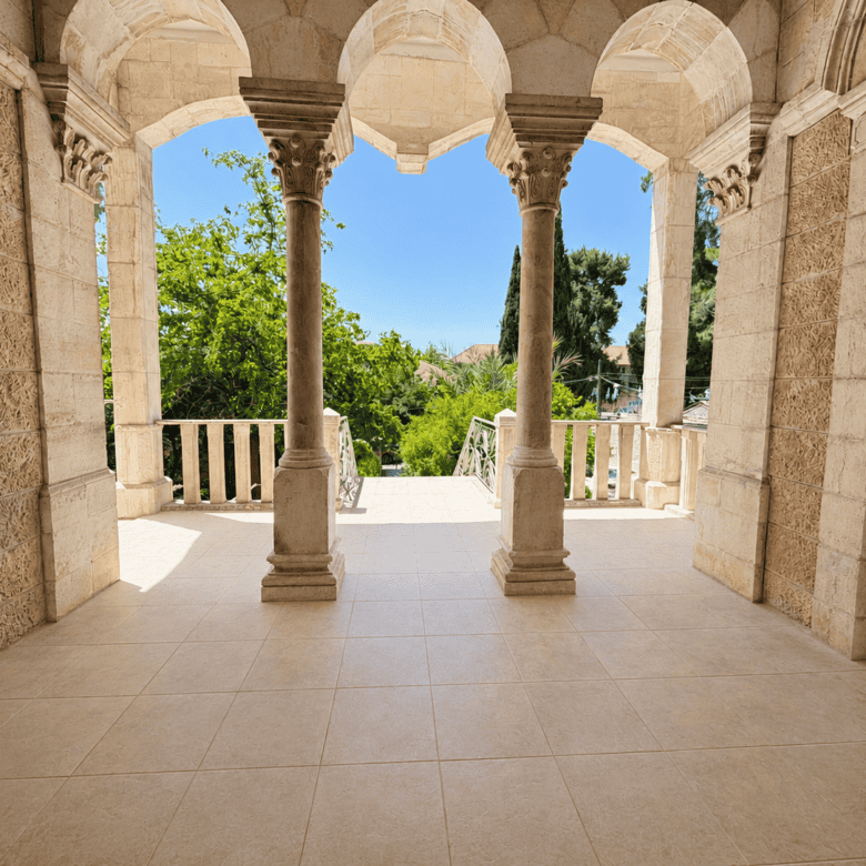 Stone arcade with two columns on a tiled balcony, opening to a sunlit garden and blue sky beyond.