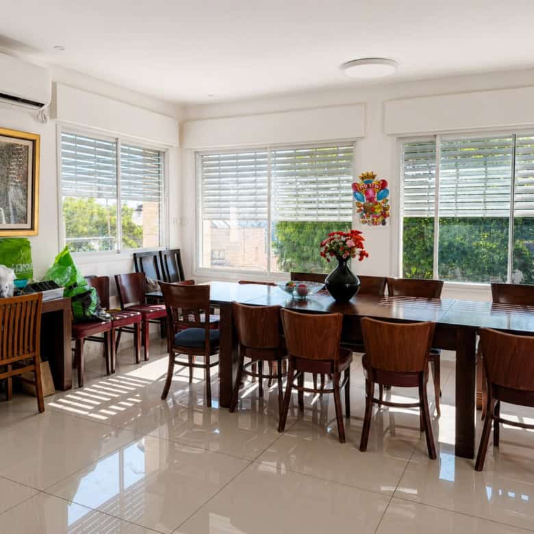 Sunlit dining room with a long wooden table and multiple chairs, large windows with white blinds, and a vase of red flowers as a centerpiece.