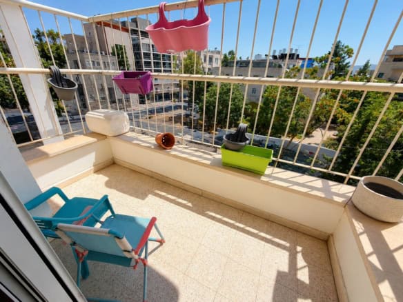 Small balcony with colorful plant pots hanging on a white railing and a blue folding chair, overlooking a city street and trees.