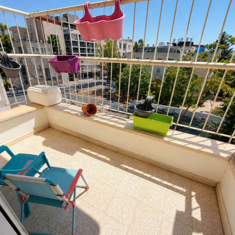 Small balcony with colorful plant pots hanging on a white railing and a blue folding chair, overlooking a city street and trees.