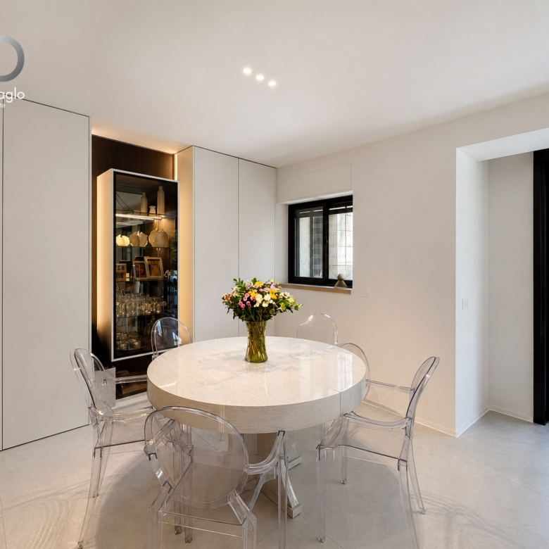 Modern dining room with a round marble table, clear acrylic chairs, and a vase of flowers; glass display cabinet along a white wall.
