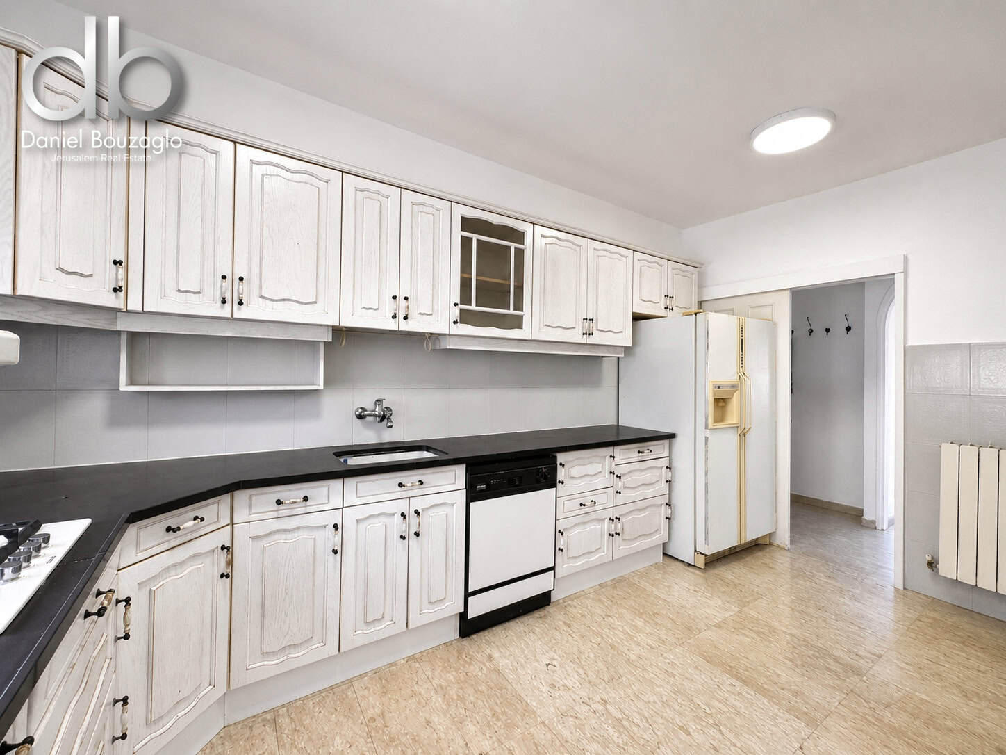 Long white kitchen with raised-panel cabinets, black countertops, a white dishwasher, and a beige fridge by a doorway.