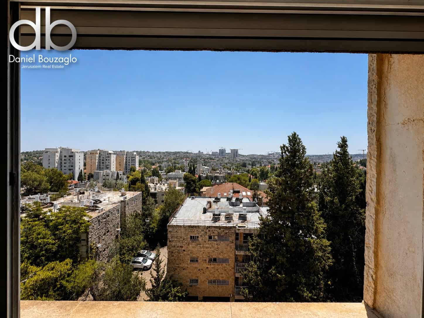 View of a sunlit cityscape from a window, with mid-rise buildings, trees, and a blue sky in the distance.