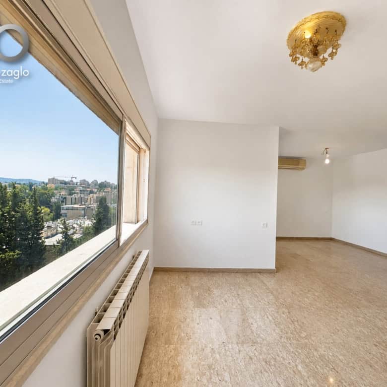 Bright empty living room with a large window showing a city view, beige floors, white walls, radiator, and ceiling light fixture; real estate watermark in corner.