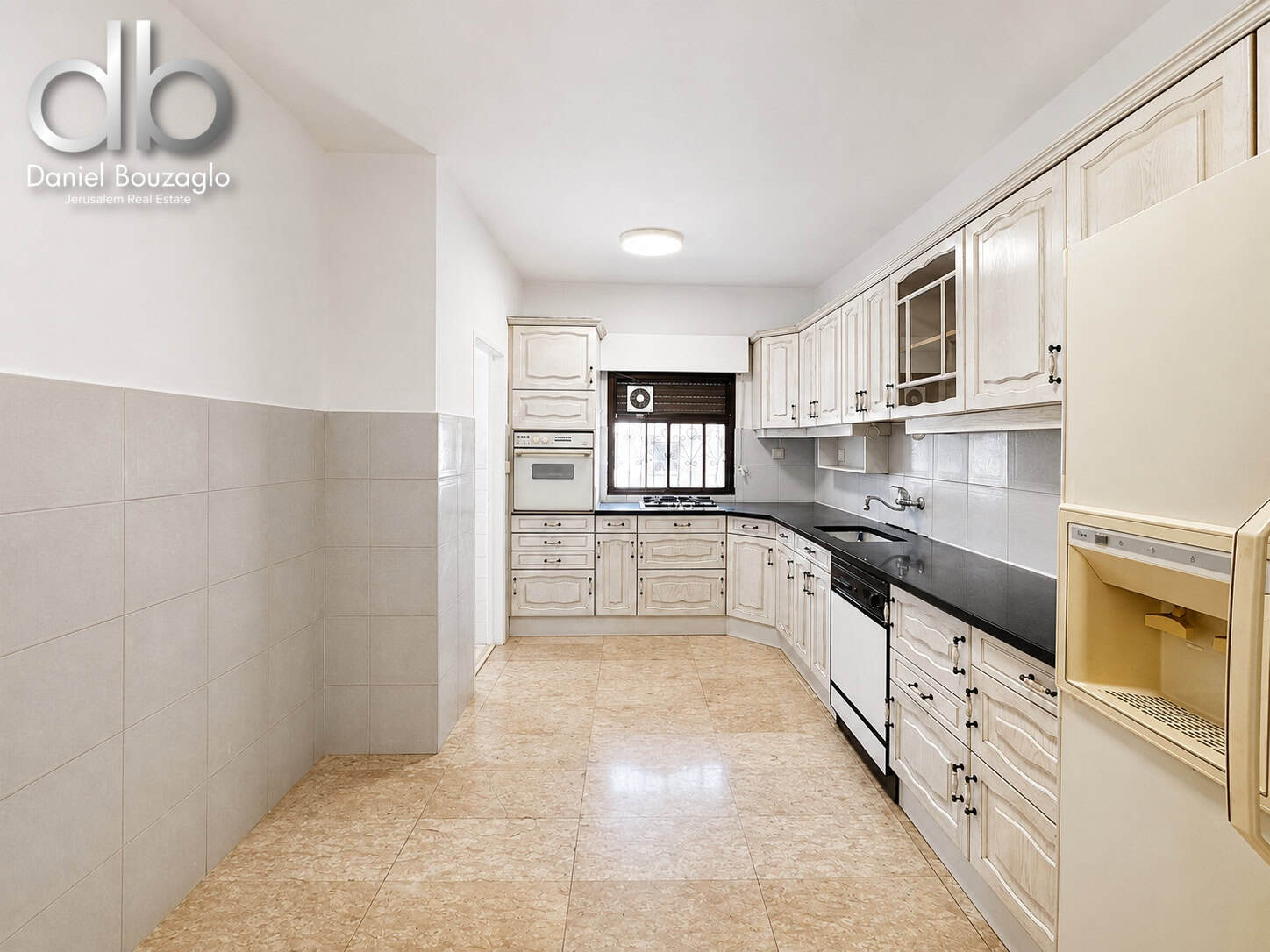 Bright L-shaped kitchen with white distressed cabinets, black countertops, gas stove, built-in oven, sink, and beige tiled floor. (Real estate photo showing a spacious, light-filled kitchen.)