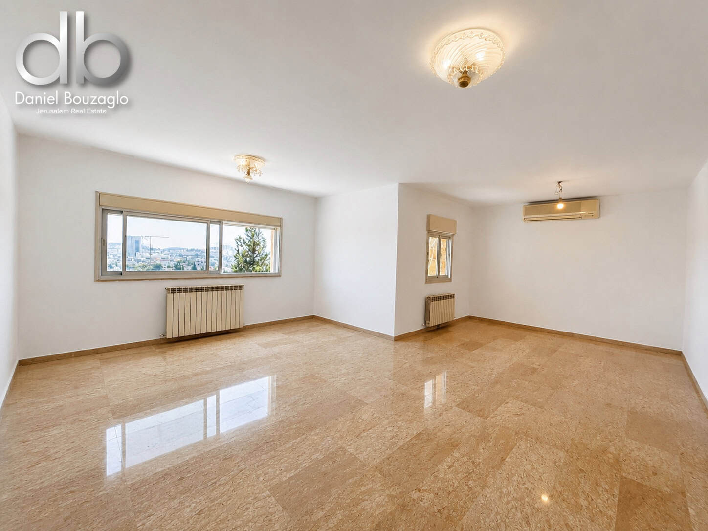 Bright, empty living room with large window, beige tiled floor, and wall radiators; logo in top-left corner.