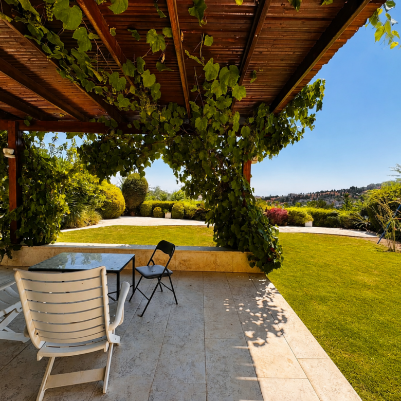Patio under a wooden pergola with green vines, white chairs, and a glass table overlooking a sunny garden lawn.