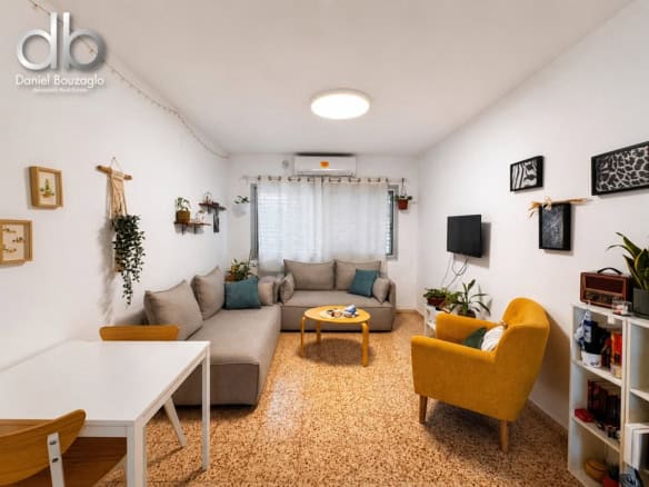 Cozy living room with an L-shaped beige sofa, mustard-yellow armchair, wooden coffee table, and several plants near a bright window. Wall art, a TV on the right, and a white shelving unit add storage and decor.