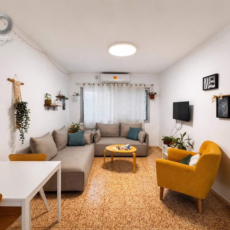 Cozy living room with an L-shaped beige sofa, mustard-yellow armchair, wooden coffee table, and several plants near a bright window. Wall art, a TV on the right, and a white shelving unit add storage and decor.