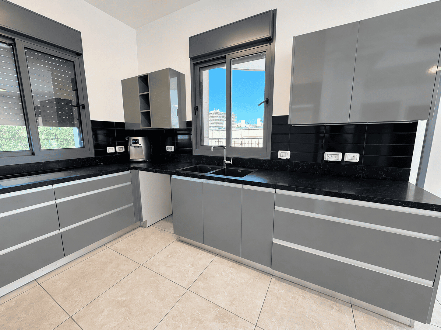 Modern L-shaped kitchen with gray cabinets, black countertop, and double sink under a window; stainless appliances nearby.