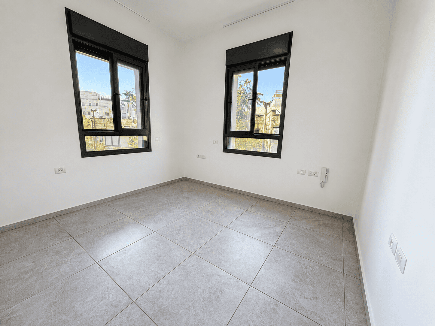 Empty bright room with two large black-framed windows, white walls, and gray tiled floor.