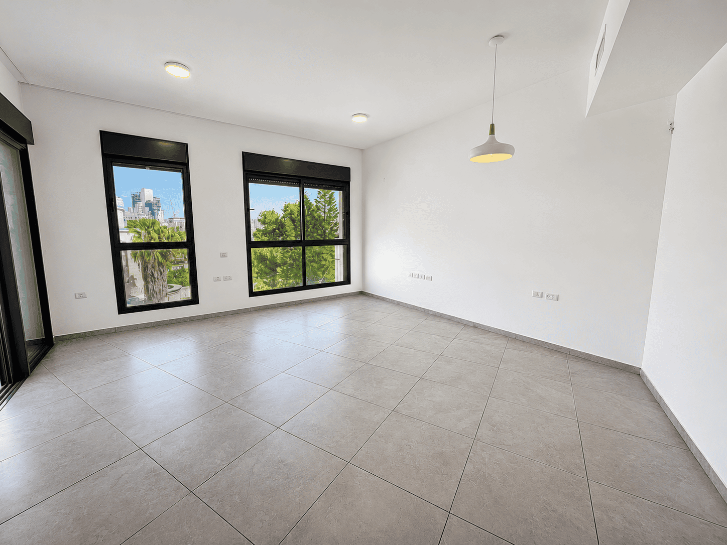 Bright empty room with large black-framed windows, white walls, and gray tiled floor; a pendant light hangs from the ceiling with a view of trees outside the windows.