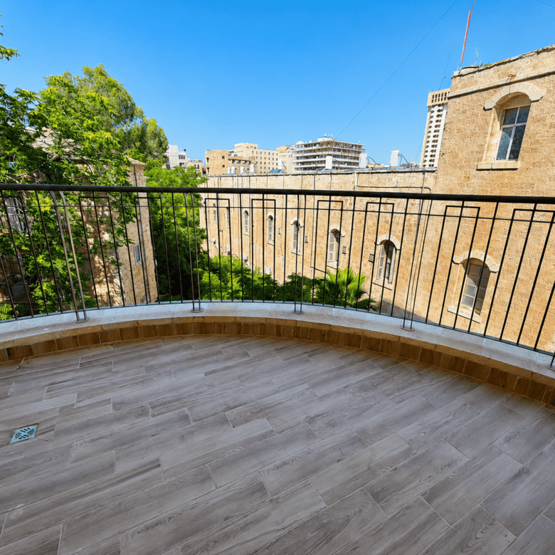 Curved balcony with a black metal railing, light wood-look tiles, overlooking sandstone buildings and green trees under a bright blue sky.